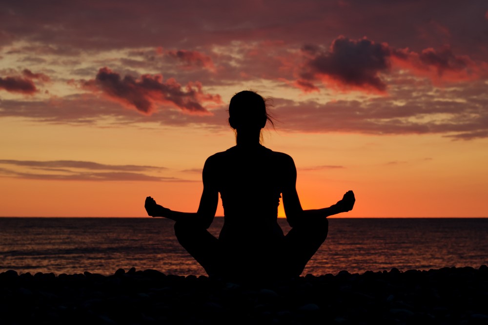 Silhueta de uma mulher meditando na praia durante o pôr do sol. A mulher está sentada em posição de lótus com as mãos em seu colo, olhos fechados. O céu está cheio de nuvens rosa e laranja.
