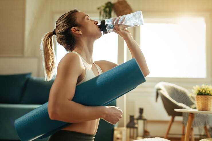 Mulher praticando exercícios em casa, segurando um tapete de yoga azul e bebendo água de uma garrafa plástica. 