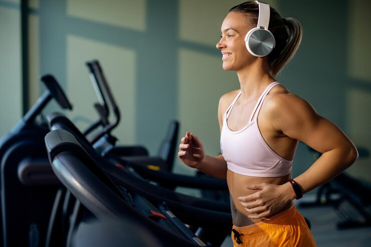 Mulher correndo em uma esteira na academia, usando fones de ouvido e sorrindo. Está aquecendo para evitar lesões comuns na musculação.