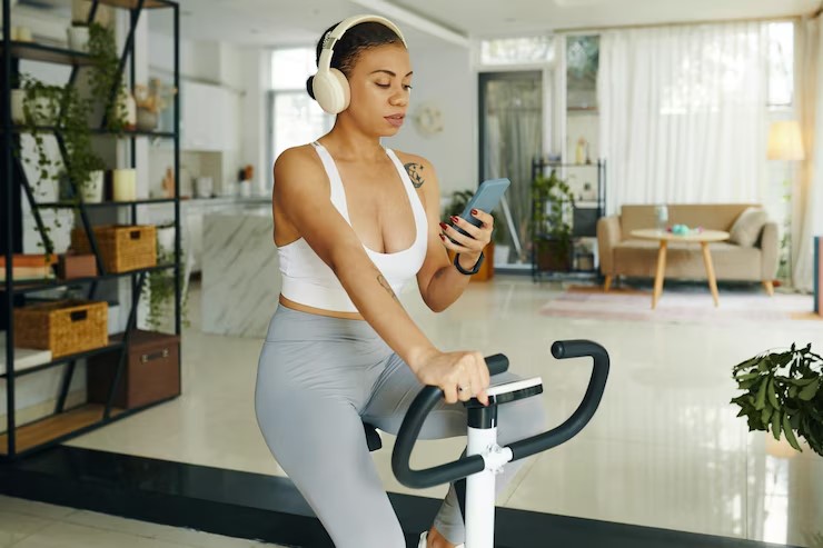 Mulher usando fones de ouvido enquanto pedala em uma bicicleta ergométrica em casa, incorporando exercícios na rotina diária.