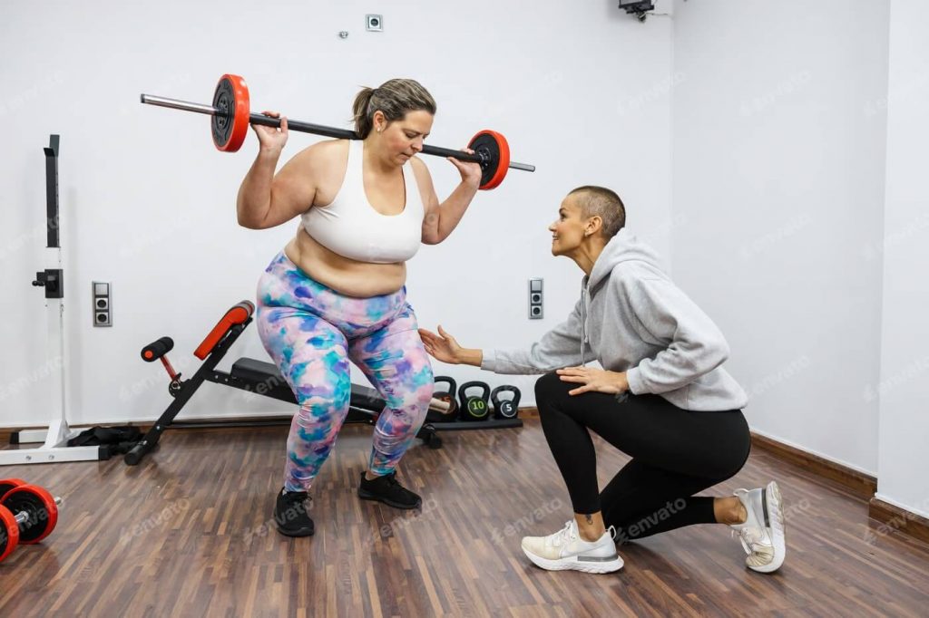 Instrutora auxiliando uma mulher a realizar o levantamento de peso da forma correta.