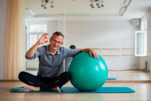 Homem em sua sala sentando em um tapete de pilates apoiado em uma bola de pilates