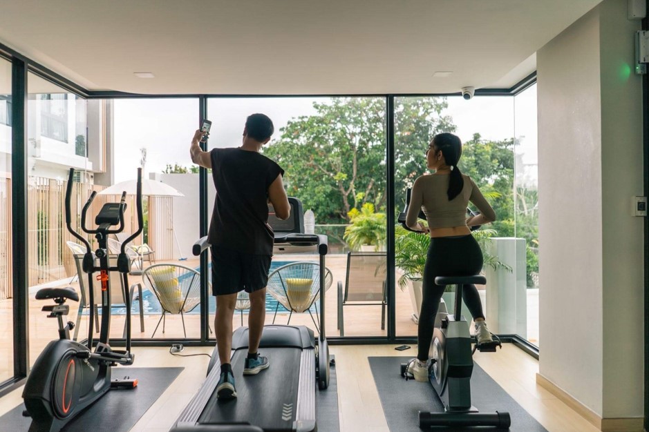 Casal se exercitando na academia com vista para a piscina; o homem caminha na esteira e a mulher usa a bicicleta ergométrica ao treinar no condomínio.