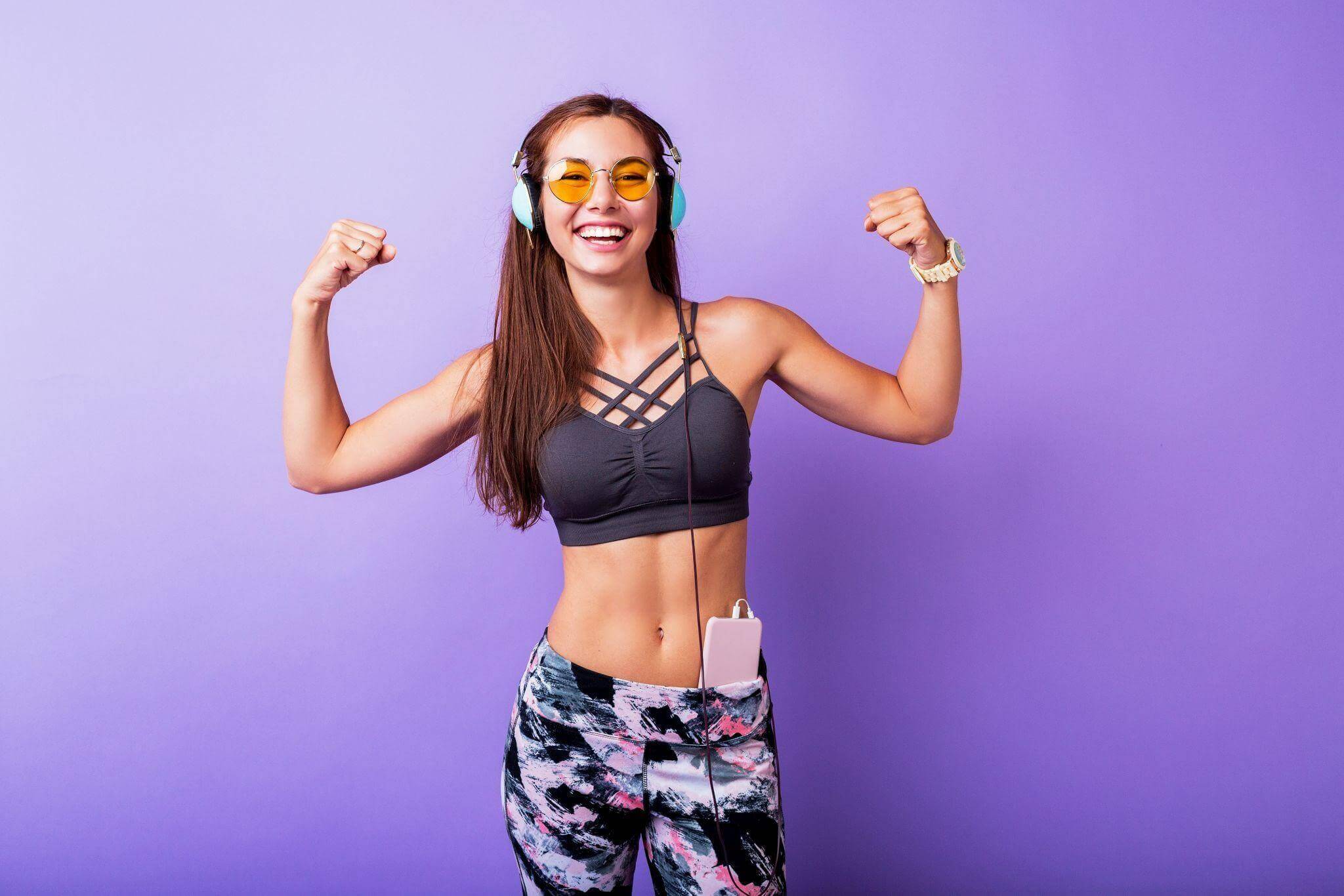 Mulher jovem de pele clara e cabelos castanhos longos sorri alegremente enquanto faz uma pose de "muque" com os dois braços. Ela usa óculos de sol redondos com lentes amarelas, fones de ouvido grandes azul-claros e um top esportivo cinza. Um celular está guardado no cós de sua calça legging estampada. O fundo é de uma cor lilás sólida e vibrante.