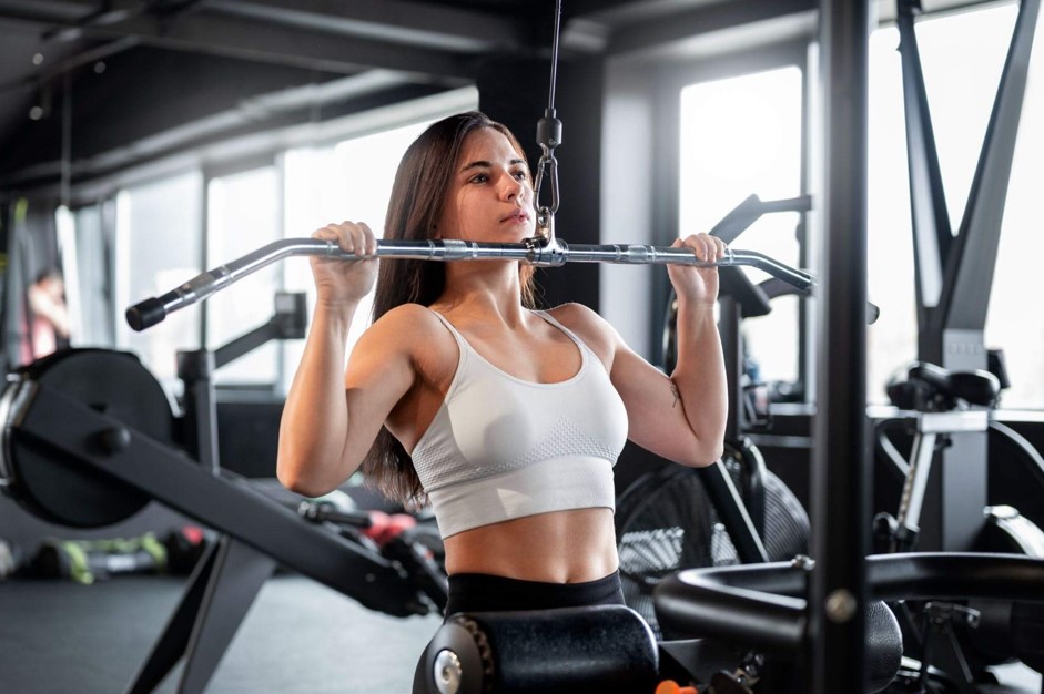 Mulher jovem treinando em um aparelho de puxada alta (pulley) para completar suas séries de musculação. Ela segura a barra com pegada aberta, demonstrando concentração e esforço muscular em uma academia bem iluminada.