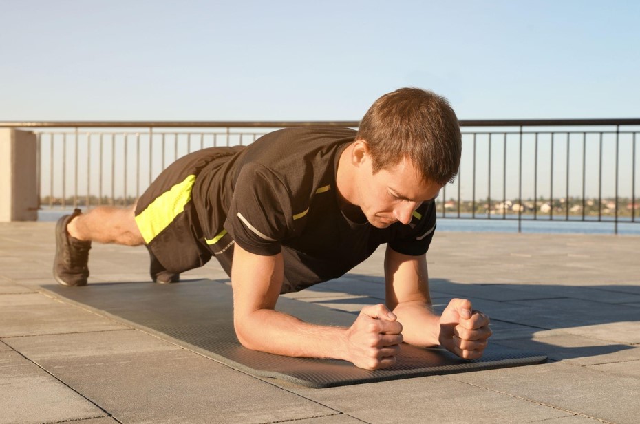 Um homem jovem realiza a prancha abdominal sobre um tapete de yoga em uma área externa pavimentada. Ele mantém o corpo alinhado e os antebraços apoiados no chão, demonstrando um dos exercícios para articulações que foca na estabilidade do core e proteção da coluna.