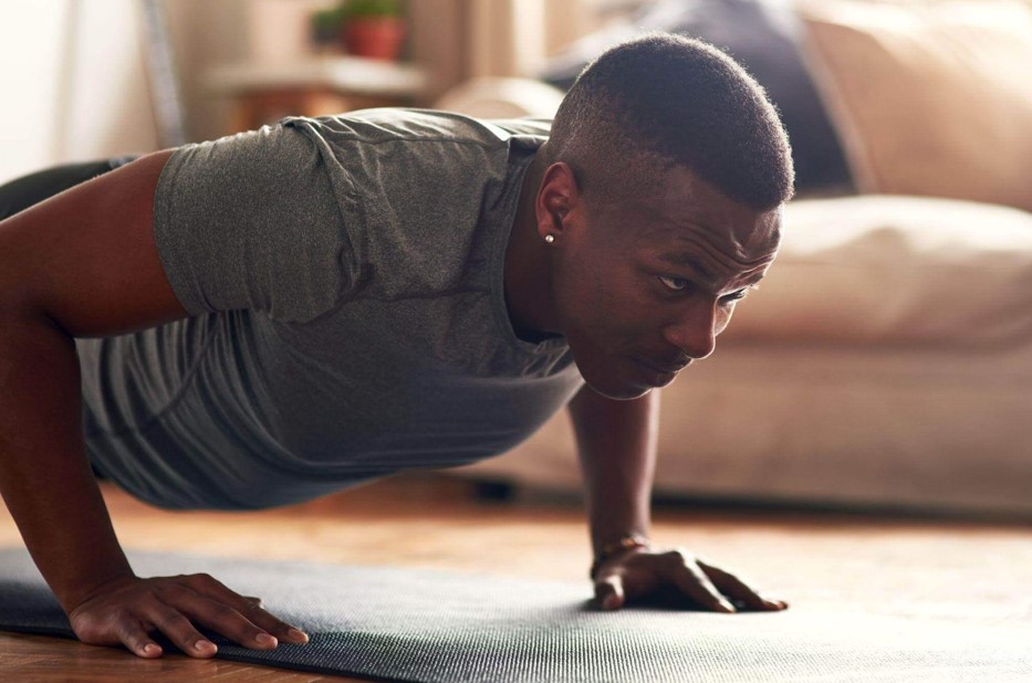 Homem negro focado realizando flexão de braços sobre um tapete de yoga em casa. Este exercício de força é um excelente complemento para treinos que incluem agachamentos para trabalhar o corpo todo.