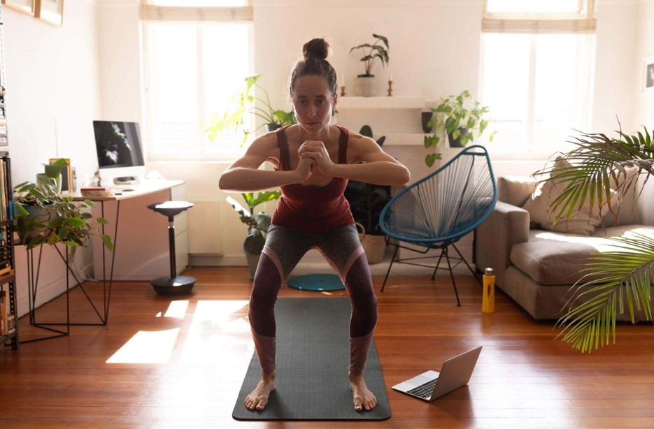 Mulher em posição inicial de agachamentos, com as mãos unidas à frente do peito, treinando na sala de casa com o auxílio de um notebook e cercada por plantas.