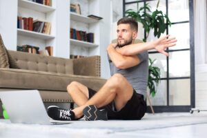Homem jovem sentado no chão de uma sala de estar, realizando um alongamento de braço cruzado sobre o peito. À sua frente, há um laptop aberto, indicando que ele segue uma rotina guiada para ajustar suas séries de musculação e flexibilidade no ambiente doméstico.