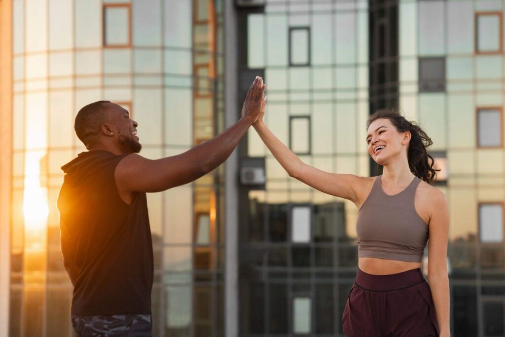 Homem e mulher sorrindo e comemorando com um "high five" após o treino ao ar livre no pôr do sol. A imagem reflete a energia e o bem-estar proporcionados pela rotina fitness, que pode ser mantida com a correta manutenção de equipamentos de academia em casa.