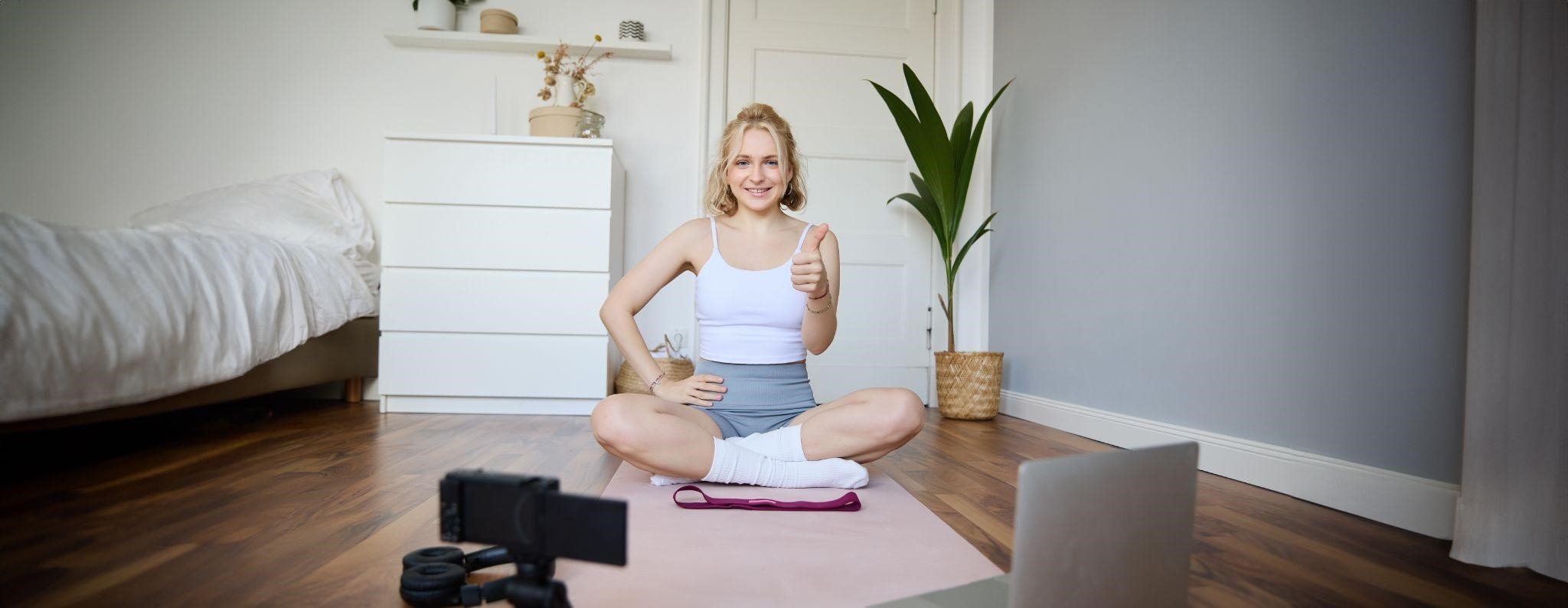 Mulher loira sorrindo sentada em pose de ioga sobre um tapete rosa, gravando um treino com o celular em um tripé e um notebook à frente. Ela faz um gesto de positivo com o polegar, mostrando como montar uma funcional academia em casa 4m² no próprio quarto, ao lado da cama.